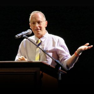 David Sedaris stands at a lectern. He smiles and is speaking into a microphone.