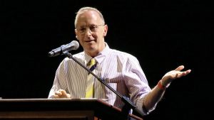 David Sedaris stands at a lectern. He smiles and is speaking into a microphone.