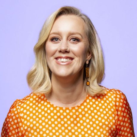 Comedian Claire Hooper smiling in front of a purple background. She wears an orange and white polka-dot dress.