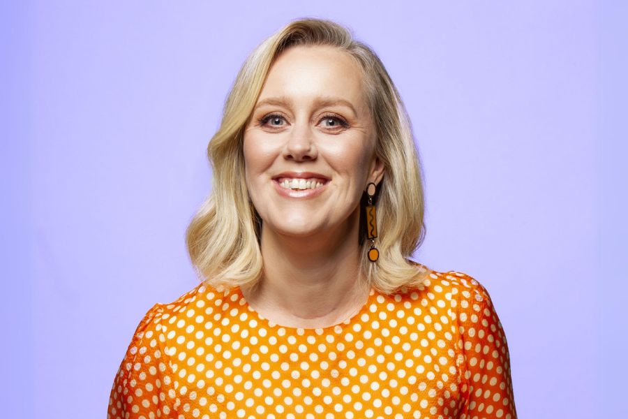 Comedian Claire Hooper smiling in front of a purple background. She wears an orange and white polka-dot dress.