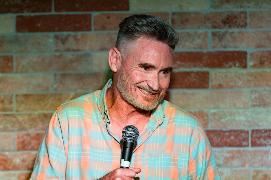 Comedian Dave Hughes smiling with a microphone in his hand. He's in front of a brick wall wearing a plaid shirt.