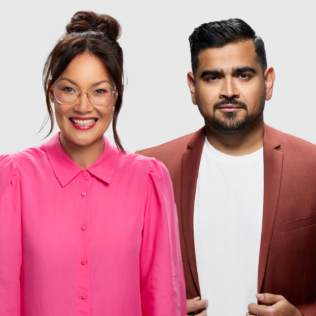 Lizzy Hoo stands in front of and to the right of Dilruk Jayasinha. Both face the camera. Lizzy is smiling brightly, her teeth showing between her red lipstick. She wears a hot pink, button-up blouse with a collar. Her black hair is bundled into a rough bun with two distinct bangs framing her face. She has big, light grey framed glasses on. Dilruk has one eyebrow raised, smoldering at the camera. He holds the bottom of the long, thin lapels of his mid-brown jacket, which he wears over a stark white t-shirt. He has a neatly trimmed, short black bread and moustache and his black hair is combed back and parted on his left. The backdrop is white.