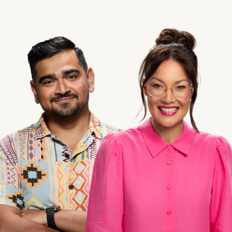 Lizzy Hoo stands in front of and to the right of Dilruk Jayasinha. Both face the camera. Lizzy is smiling brightly, her teeth showing between her red lipstick. She wears a hot pink, button-up blouse with a collar. Her black hair is bundled into a rough bun with two distinct bangs framing her face. She has big, light grey framed glasses on. Dilruk smiles gently with his head tilted to the left. He wears a short sleeved shirt with various patterns in many pastel colours. He has a neatly trimmed, short black bread and moustache and his black hair is combed back and parted on his left. The backdrop is off white.