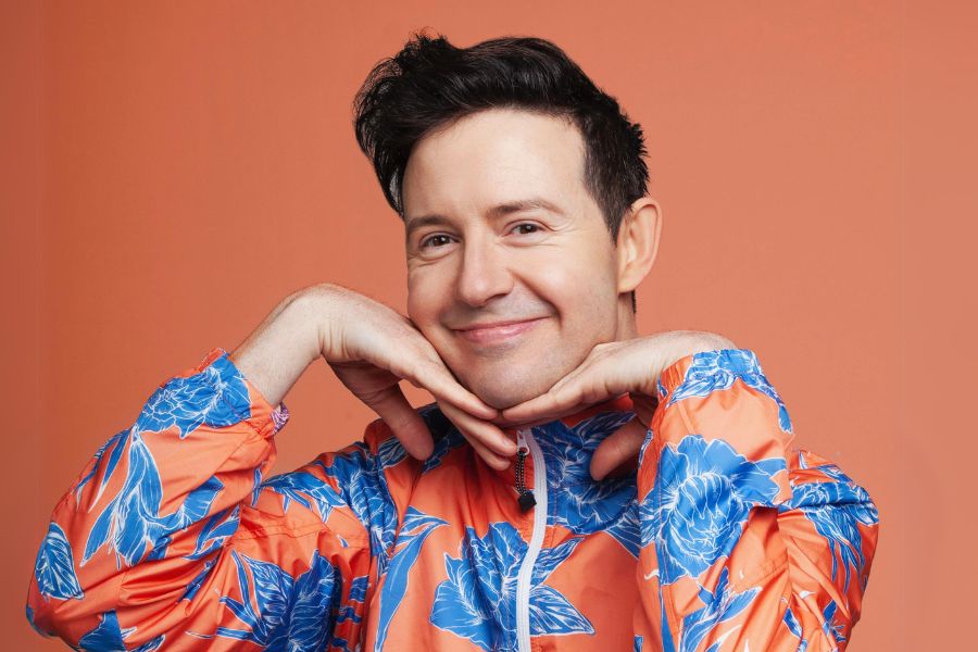 Comedian Nath Valvo smiling cheekily in front of an orange background. He wears a blue and orange puffer jacket.