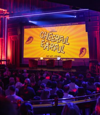 A stage with a large screen reading 'Cheeful Earful'. A large live audience.