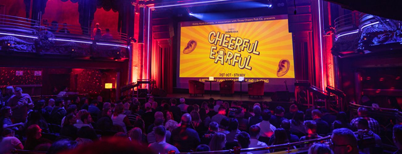 A stage with a large screen reading 'Cheeful Earful'. A large live audience.