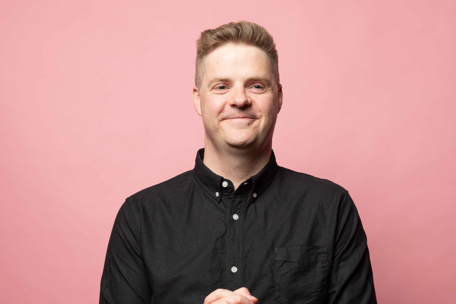 Tom Ballard has short blonde hair and blue eyes. He is wearing a black buttoned up top and is standing in front of a light pink backdrop. He is smiling just outside of the camera lens and has his hands clasped together in front of his stomach. 