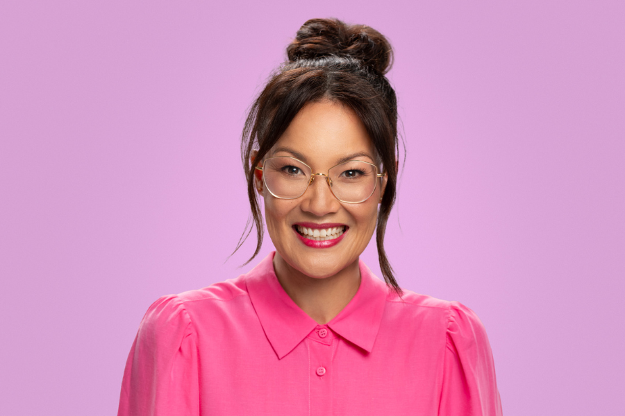 Lizzy Hoo has dark brown hair in a top bun. She is wearing big round glasses and a pink button up shirt. Lizzy is smiling directly into the camera. She is in front of a purple backdrop. 