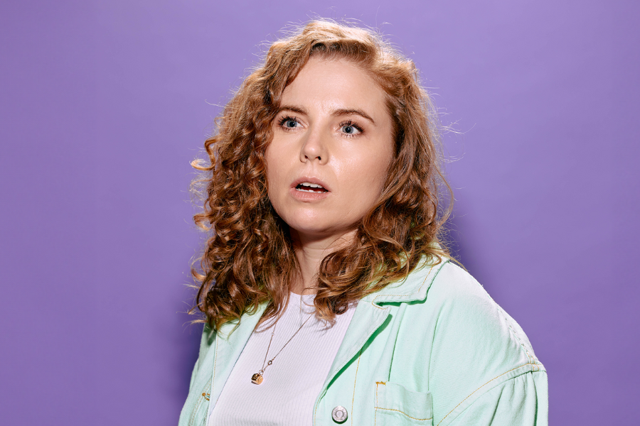 Bronwyn Kuss has long strawberry blonde curly hair. She is wearing a white shirt with a mint green button up. Her mouth is gaped and she is looking out of camera. The background is light purple. 