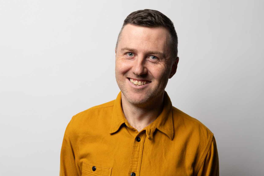 Lloyd Langford looks directly at the camera, smiling generously, his body tilted slightly to his left. His short, brown hair is neatly combed and he has very short stubble on his face. He wears a mid-to-dark yellow button up jacket with breast pockets on each side. The background is light grey. 