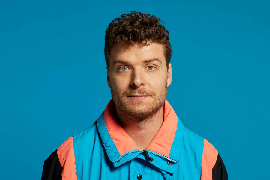 Oliver Coleman wears a bright blue and orange jacket. He has short curly hair and blue eyes. He is smirking into the camera lense.