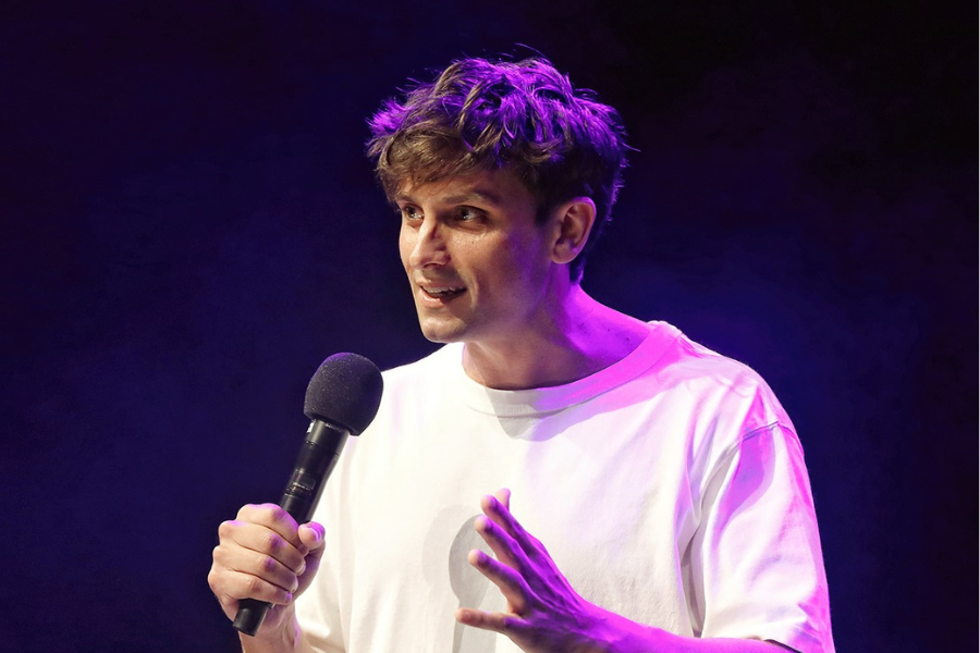 Image is of Sam Taunton on stage holding a microphone. He has short brown hair, brown eyes and is wearing an oversized white t-shirt. A purple light is hitting the back of his head, which is changing the colour of his shirt and hair. The background is dark blue. 