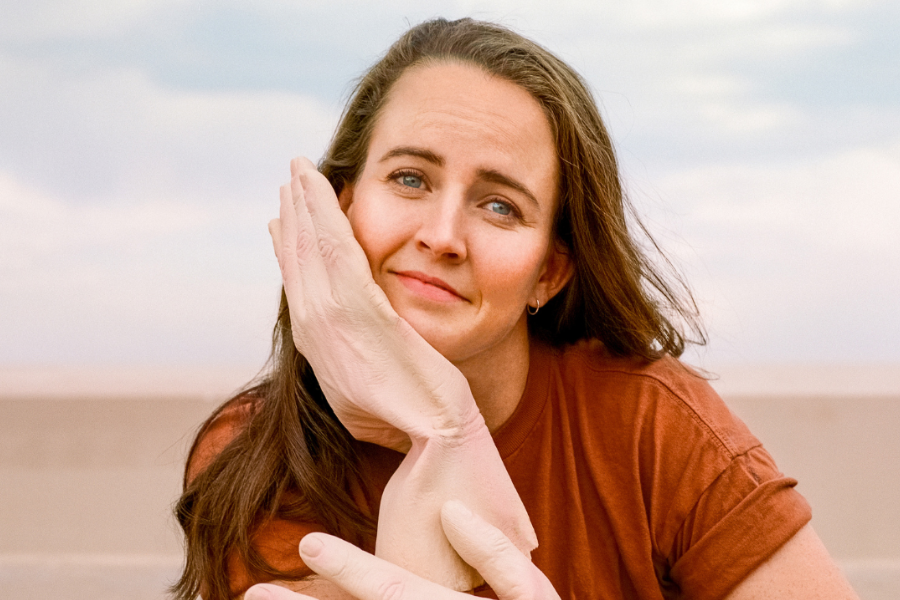 Zoë Coombs Marr has long brown hair and blue eyes. She is wearing a burnt orange t-shirt and oversized novelty rubber gloves that look like hands. The background is a blue sky and the foreground is a dark wooden table. She is smiling at the camera. 