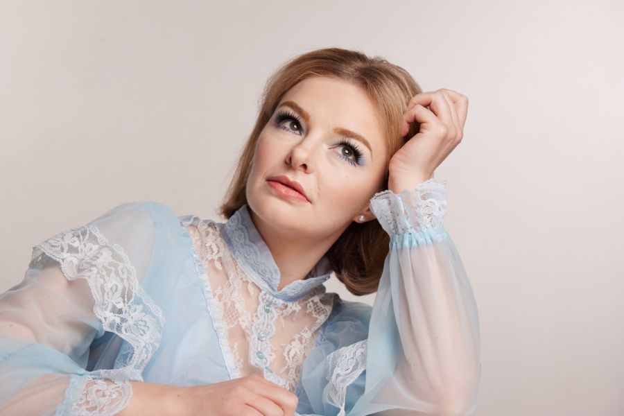 Image is of Abby Howells. She has blue eyes and blonde hair that is cut into a bob. She is wearing a light blue lacy dress and pearl white earrings. He right arm is bent and her head is leaning on her left hand. She is looking upwards.