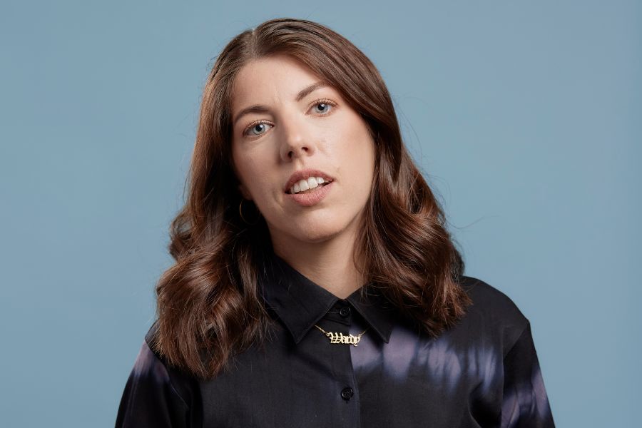 Alex Ward stands front on to the camera, her head tiled gently to her right. She smiles gently with her teeth showing and her blue eyes look directly and warmly to the camera. Her long brown hair drops below her shoulders. Her button up long sleeved shirt is black silk with the odd row of soft white tie die blurs. Between her collar ends is a fine gold chain with the word Wardy in stately lettering. The background is a grey blue.  