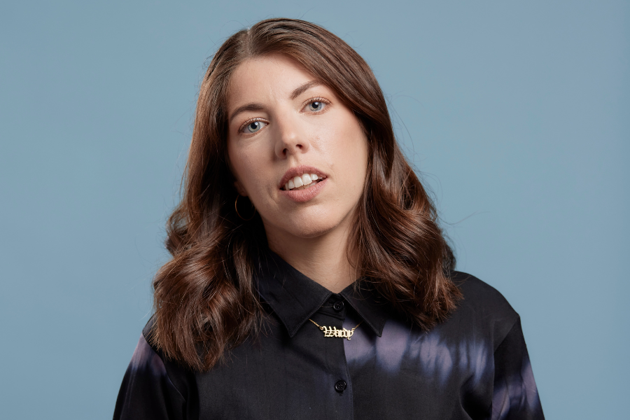 Alex Ward stands front on to the camera, her head tiled gently to her right. She smiles gently with her teeth showing and her blue eyes look directly and warmly to the camera. Her long brown hair drops below her shoulders. Her button up long sleeved shirt is black silk with the odd row of soft white tie die blurs. Between her collar ends is a fine gold chain with the word Wardy in stately lettering. The background is a grey blue. 