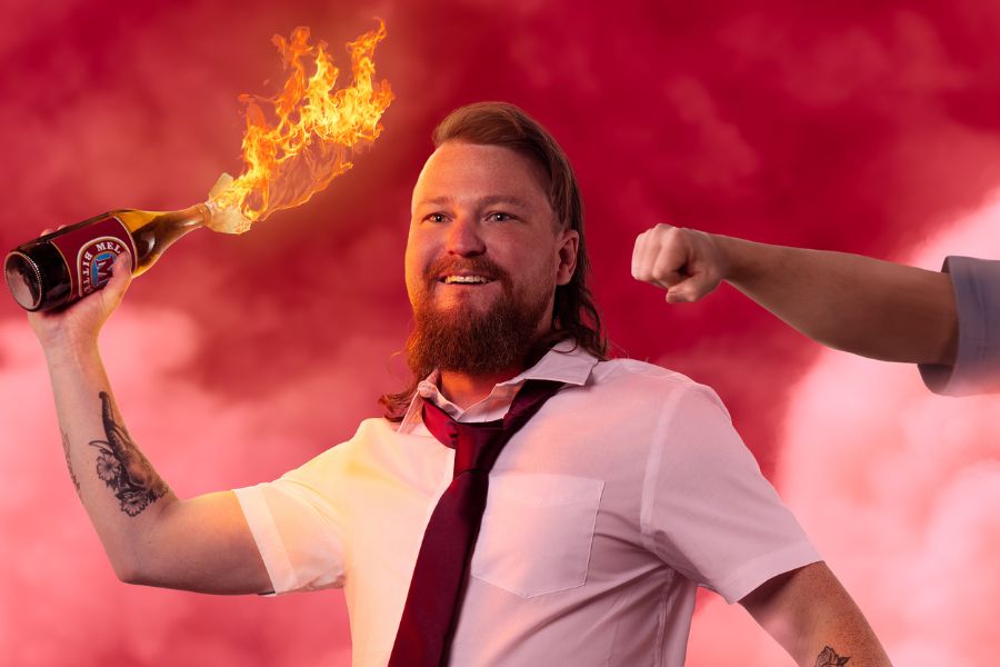 Brett Blake stands confidently in front of swirling red smoke, wearing a white button-up shirt and a red tie. In his hand, a beer bottle is ablaze, casting an orange glow in the chaotic scene. A fist rushes into the frame from the right, suggesting an impending clash. The fiery, tumultuous backdrop contrasts sharply with Brett’s composed appearance, creating a tense, action-packed atmosphere.