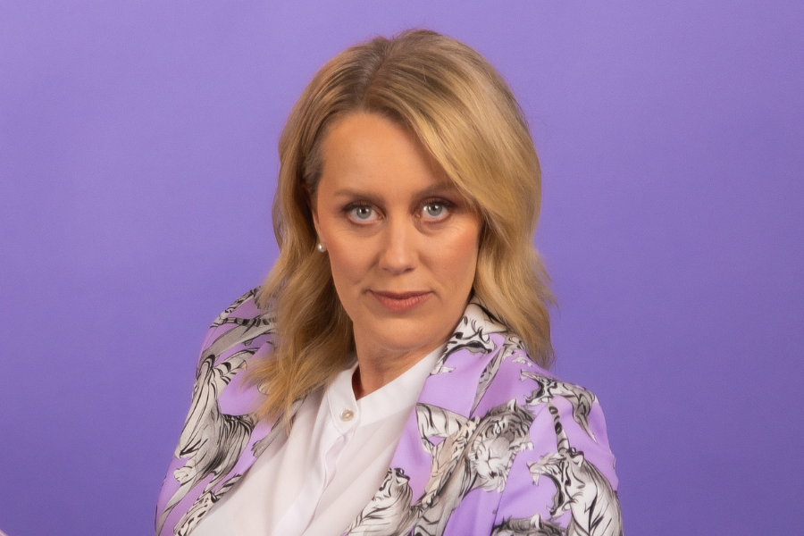 Claire Hooper faces the camera with a neutral expression on her face and her body turned to her right. She is to the right of frame, so there is space to the left of frame. Her gently wavy, champagne blonde hair drops below her shoulders. She wears a lilac suit with black & white tigers on it and a white dress shirt. The background is purple. 