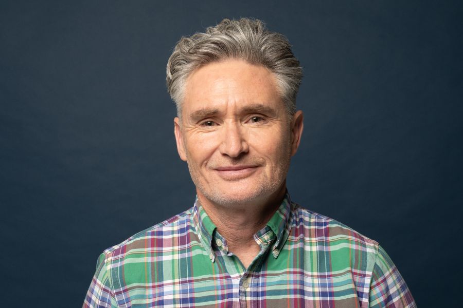 Image is of Dave Hughes. He is wearing a green, red & blue checked shirt. He is looking towards the camera. He has short wavy grey hair and stubble. The background is navy blue.