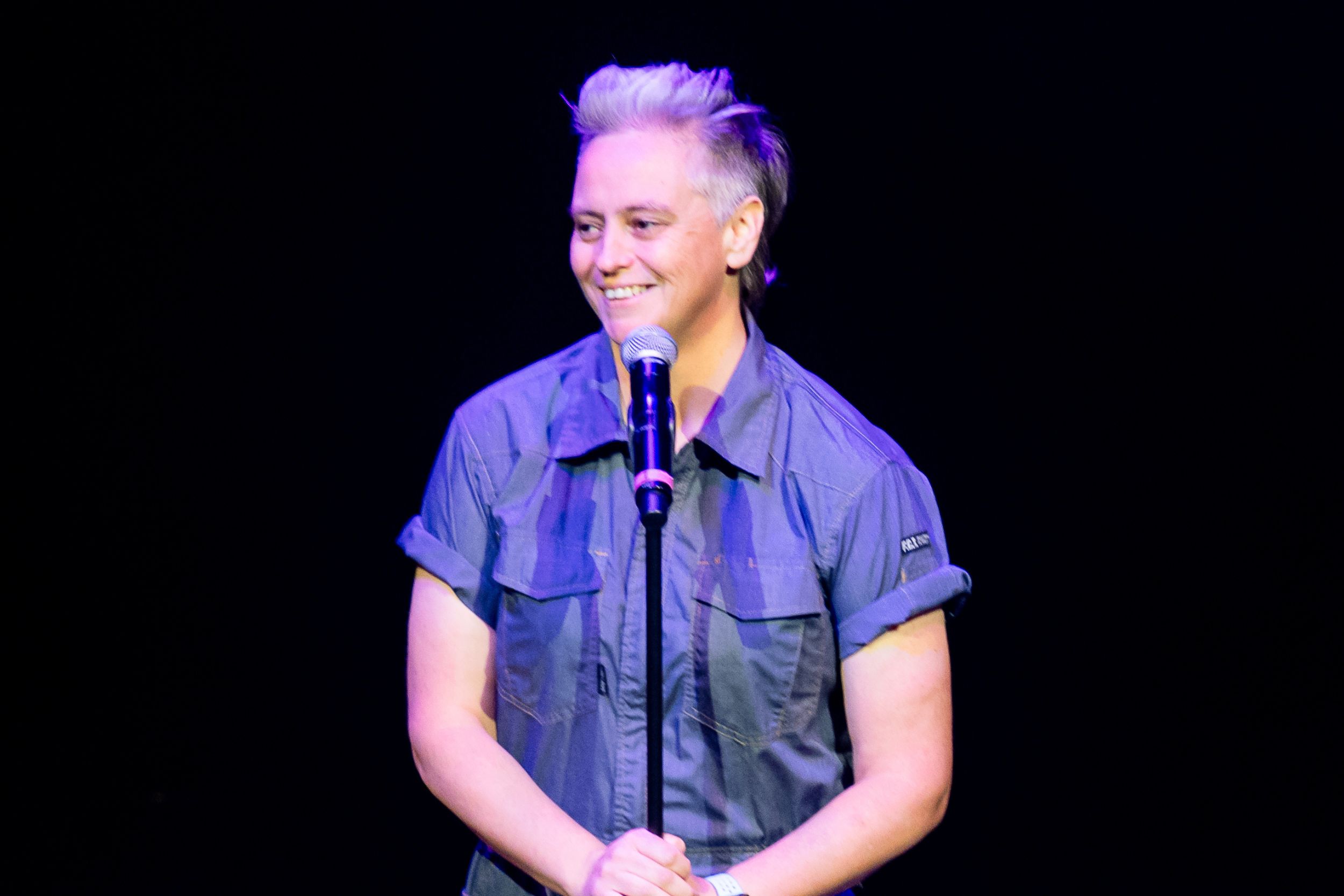 Image is of Geraldine Hickey wearing a blue boiler suit. She is performing stand up comedy.