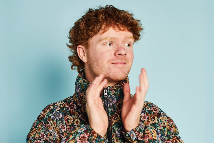 Image is of Will Gibb. Will has short red curly hair. He is wearing a large puffer jacket that has a rainbow speckle pattern. He is looking out of frame and smiling, while clapping his hands. 