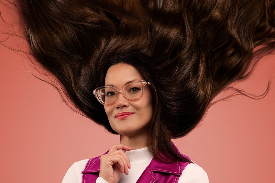 Lizzy Hoo smiles gently to the camera as her voluminous brown hair flies upwards in a V. She is turned slightly to her right. She wears a crisp white skivvy under a pink satin V neck topped dress with one large white button on it. She has large, translucent pink framed glasses on. Her right hand is up and almost closed, except for her index finger which sits above the others as if she is about to place it on her chin. The background is orange.