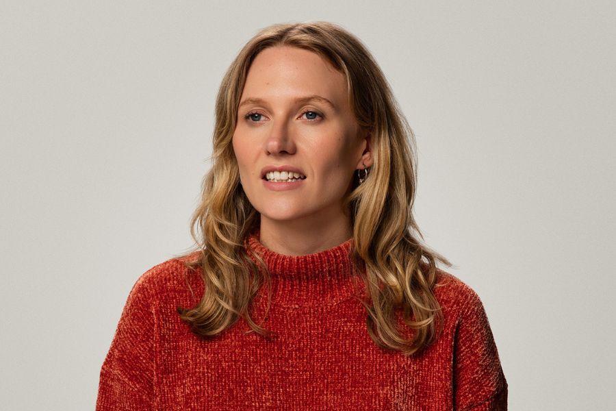 Jordan Barr looks to the left of shot with the hint of a smile on her face, her teeth showing between her open lips. Her slightly wavy, blonde hair falls just beyond her shoulders. She wears a reddish orange knitted jumper with white flecks through it and ribbing all the way round the neck. She also wears hooped earrings with a bauble at the bottom. The background is off white.