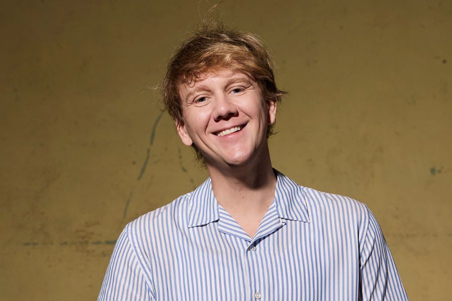 Image is of Josh Thomas wearing a blue and white stripy shirt smiling into the camera. Josh has blonde floppy hair and is standing in front of a green wall.