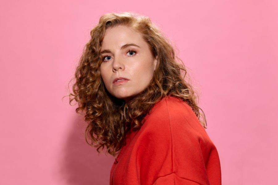 Bronwyn Kuss has her side to the camera, facing to her right, with her head turned to the camera. Her long, curly, strawberry blonde hair cascades over her shoulder. Her expression is quite neutral. She wears an orange jumper. She casts a shadow onto the baby pink background behind her. 