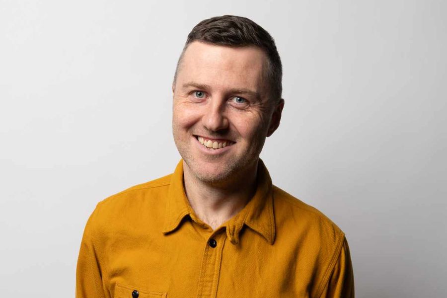 Lloyd Langford looks directly at the camera, smiling generously, his body tilted slightly to his left. His short, brown hair is neatly combed and he has very short stubble on his face. He wears a mustard yellow button up jacket with breast pockets on each side. The background is off white.