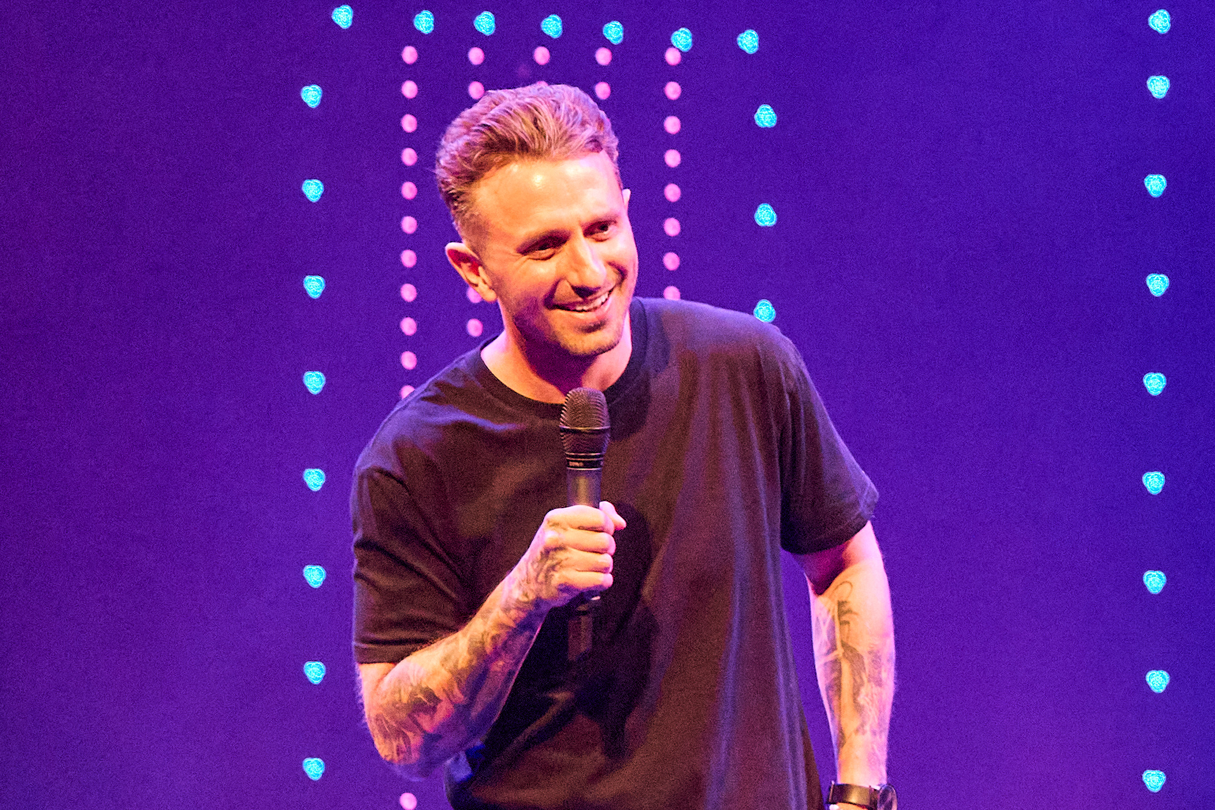 Image is of Tommy Little. He is wearing a black t-shirt and holding a microphone. He is standing on stage in front of LED lights.