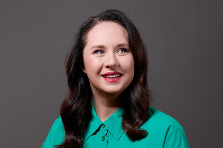 Mel Buttle has long brown wavy hair, which falls below her shoulders and red lipstick. She has a playful, bright smile as she looks to her left, her body turned a little to her right. She wears a mid-green cotton shirt. The background is chocolate brown.  