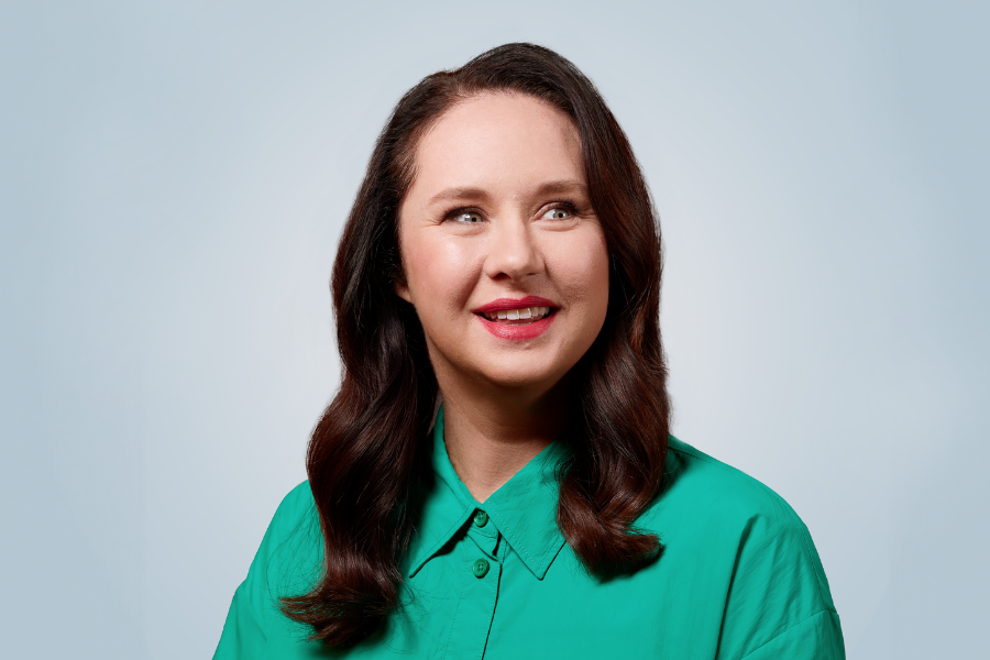 Image is of Mel Buttle wearing a teal button up shirt. She has medium length brown hair and is sitting smiling in front of a light blue background.