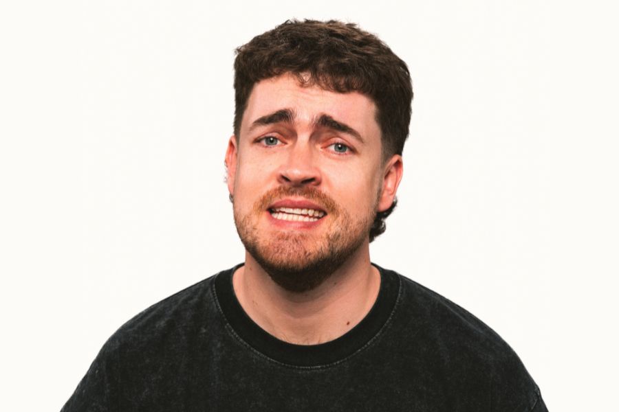 Nick White looks directly into the camera lens with an anxious look on his face, his eyebrows raised and his lips apart, the corners of his mouth pulled back to reveal his teeth. He has green eyes, short brown hair and quite pronounced stubble. He wears a dark grey t-shirt with lighter grey speckles through it and black ribbing around the neck. The background is white.