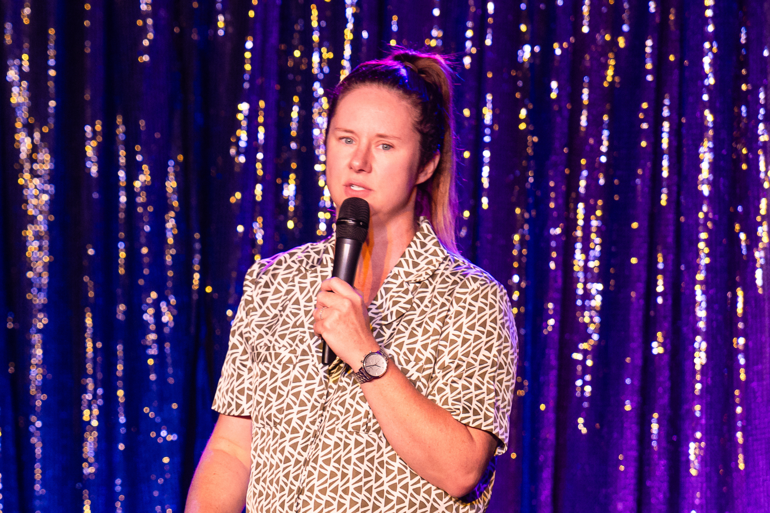 Image is of Mel Buttle performing stand up comedy. She has long brown hair which has been pulled into a ponytail. She is wearing a matching brown and white patterned set and is standing in front of a glitter curtain.