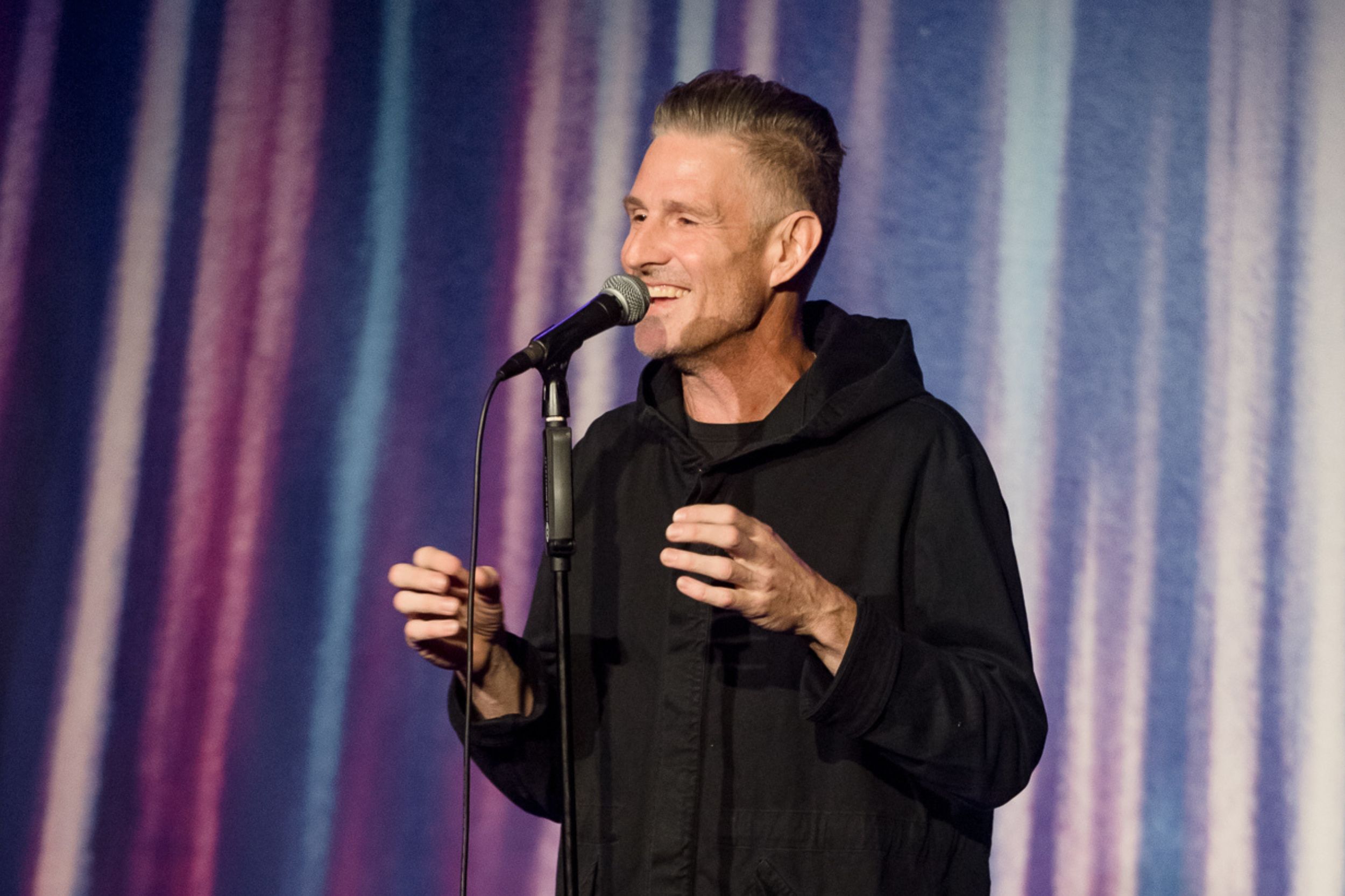 Image is of Wil Anderson performing stand up comedy. He is wearing a black jacket.