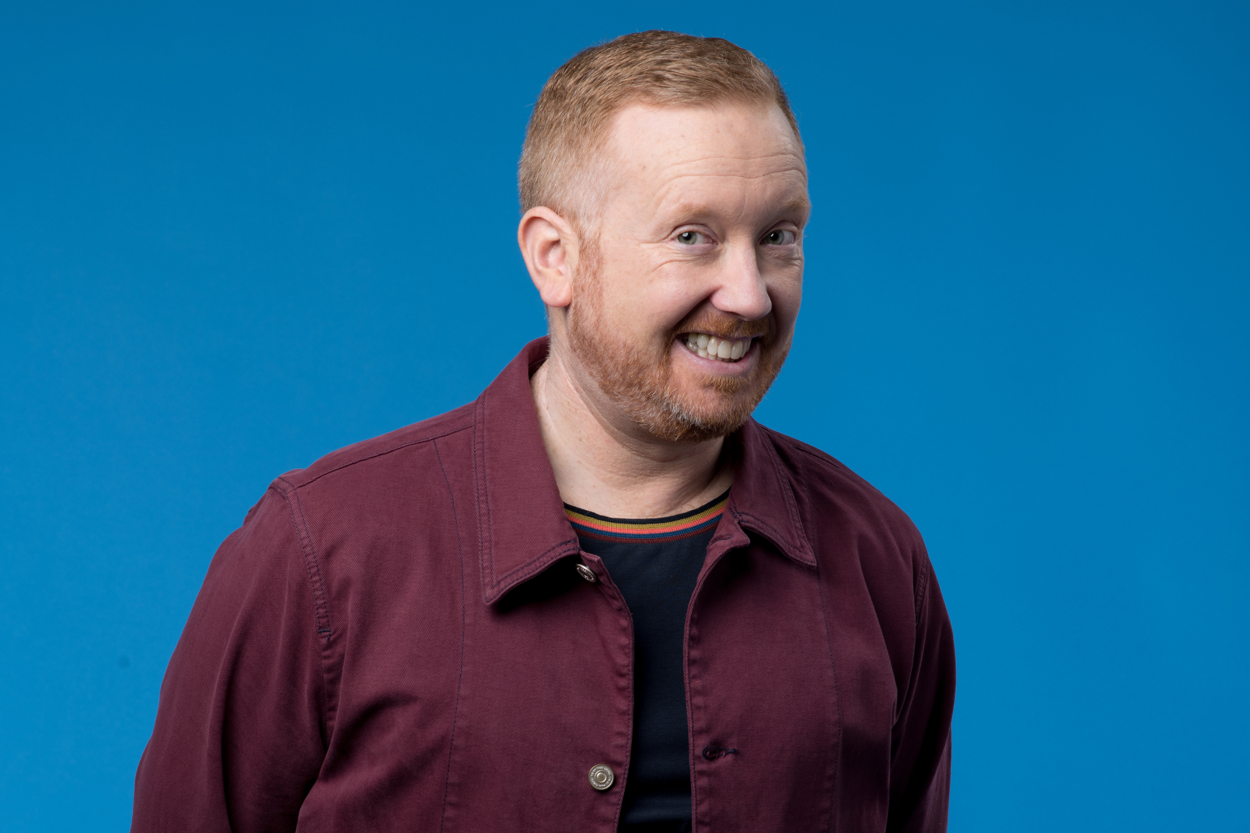 Luke McGregor has short red hair and a beard. He is standing in front of a blue background and is wearing a t-shirt with a maroon button up over the top. He is smiling with his teeth and looking into the camera.