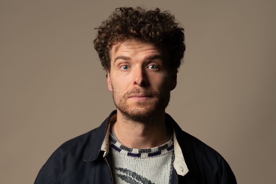 Oliver Coleman is front on to the camera looking directly into it, his grey eyes wide and his lips shut. He has curly, brown hair, a short beard, moustache and stubble. He wears a dark blue zip-up jacket over a light grey knitted jumper with a geometric design in mid and dark grey. The lighting is such that a shadow falls on the left of his face and neck. The background is fawn.