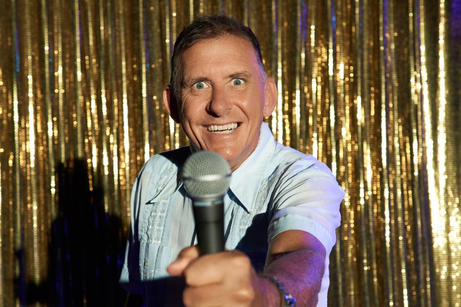 Pete Rowsthorn has a manic smile as he stares face on into the camera lens, stiffly holding a microphone up to the camera. He has a light blue, short sleeve shirt on which is textured. Behind him is a curtain of thin gold strips, some of which catch the light and reflect a bright white. He casts a heavy shadow to his right.