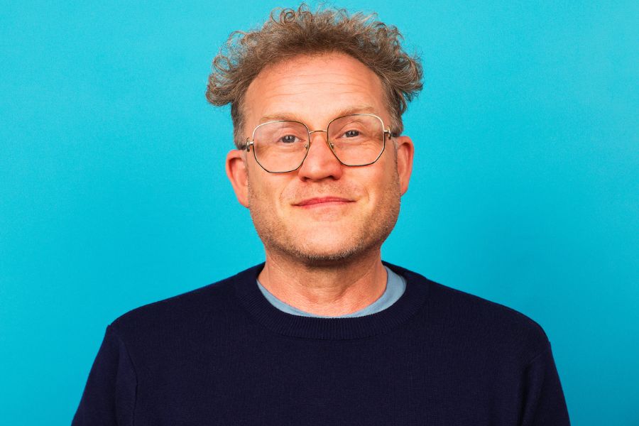 A man wearing a navy jumper with grey, red, and white stripes across the chest. He has short, light brown hair on the sides and curly hair on top. He is smiling directly at the camera, wearing large silver-framed glasses.    