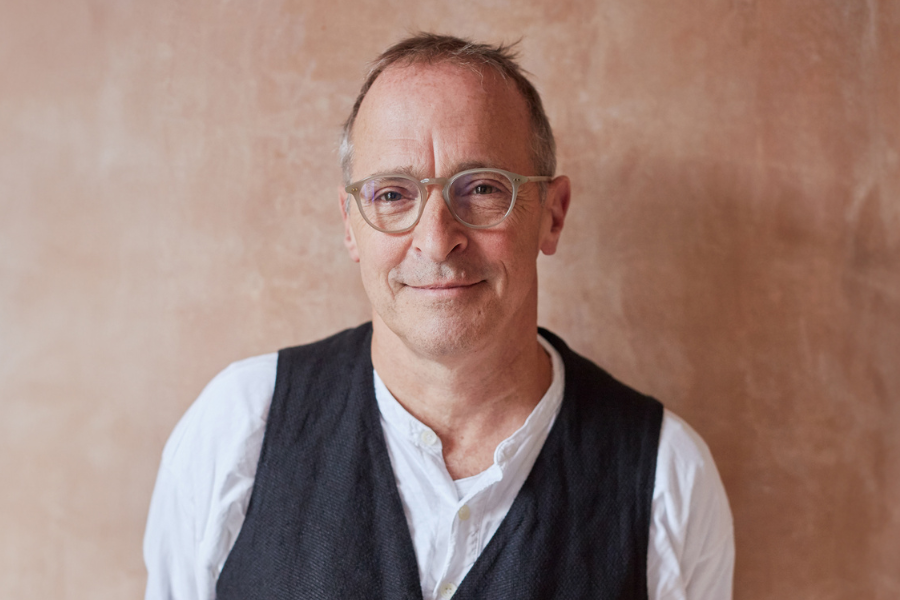 David Sedaris has wispy mid-brown hair with greying side burns. His eye glasses have light brown frames and he is smiling warmly at the camera. He has a white shirt with the top two buttons undone, under a charcoal grey vest. He is in front of a textured, caramel coloured background. 