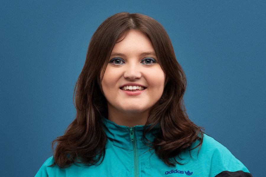 Sez looks directly to the camera, smiling generously, her top row of teeth showing. She has blue eyes and blue eye shadow on. Her wavey brown hair falls below her shoulders. She wears a turquoise green, zipped up track suit jacket which has black, purple and blue stripes across it. The background is mid-blue.