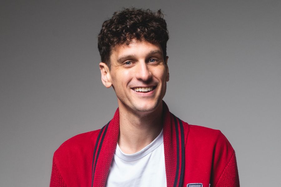 Steen Raskopoulos smiles welcomingly to camera, his head turned slightly to his left, his gaze direct into the lens. He has curly dark brown hair with short sides, blue eyes and he is clean shaven. He wears a white t shirt under a red cardigan with dark blue trim around the collar and continuing down the open front, with a light grey H on the left breast. The cardigan is much like the 1960’s fraternity house ones. The background is mid-grey.