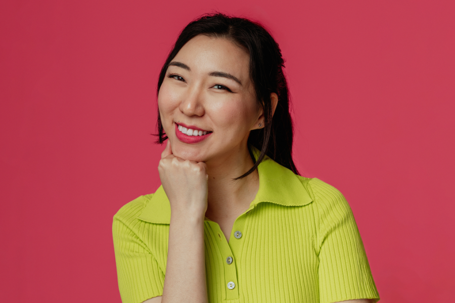 Jenny Tian smiles brightly to the camera with her right hand in a fist and her chin resting on her knuckles, her head gently tiled to her left. Her black, long straight hair is brushed back with two bangs dropping past each ear. She has red lipstick on and small silver ear studs in the shape of a simple flower. She wears a retro lime green nylon collared shirt with vertical textured lines and three buttons, the top one undone. The background is a light maroon. 