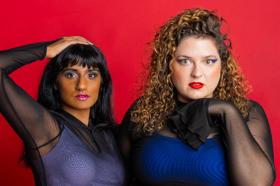 Image is of Vidya (left) and Mel (right). Both are wearing bright blue singlets with a black mesh long sleeve over the top. Vidya has dark hair and a fringe and Mel has light curly hair. The background is red.