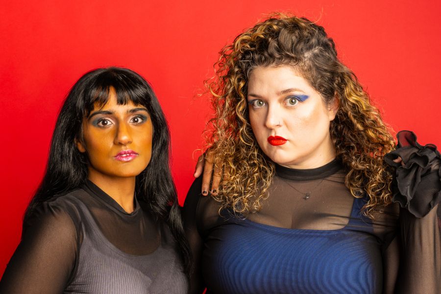 Mel (right) has long brown curly hair and Vidya (left) has short brown hair and a fringe. Both women are looking into the camera. They are standing in front of a red backdrop and wearing black mesh tops. 