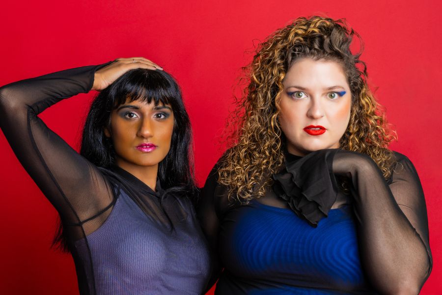 Mel (right) has long brown curly hair and Vidya (left) has short brown hair and a fringe. Both women are looking into the camera. They are standing in front of a red backdrop and wearing black mesh tops. 