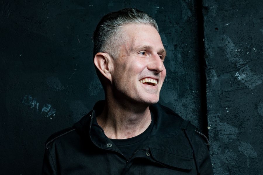 Wil Anderson has short grey hair. He is wearing a black shirt and jacket. He is looking to his right and smiling out of frame. The background is a textured black.