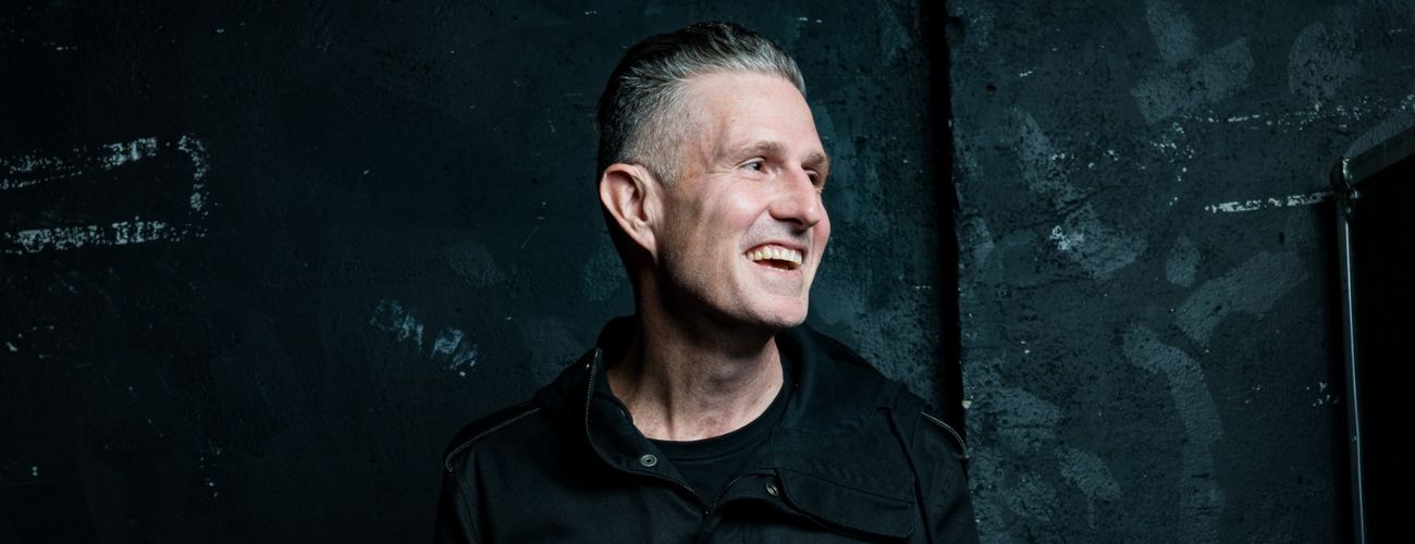 Wil Anderson has short grey hair. He is wearing a black shirt and jacket. He is looking to his right and smiling out of frame. The background is a textured black.