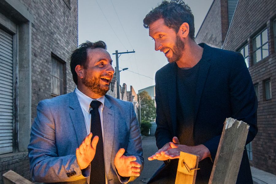 Dan Ilic and Lewis Hobba are standing in a back street, smiling at each other as they warm their hands on a fire which is burning inside a petrol drum. There are a few different lengths of wood sitting in the drum, poking up from the flames. The brick walls of the buildings on either side of the street run quite a way beyond where they stand. In the distance, in the gap between the walls, we can see blue sky. 